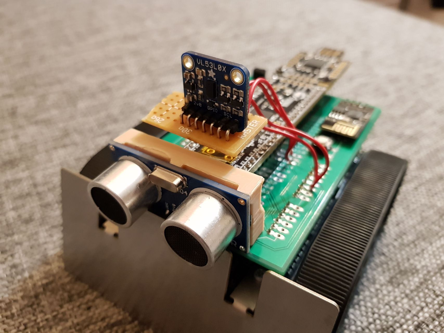 Small tracked robot with a green PCB, microcontroller and ESP8266. On the front there is an ultrasonic and IR distance sensors together with an aluminium plate for protection.