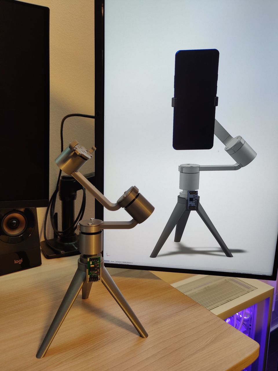 A smartphone gimbal 3D-printed out of gray plastic on a tripod. Gimbal stands on a table before a monitor displaying a CAD model of the gimbal.