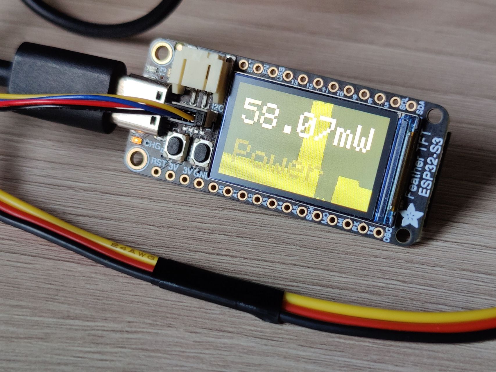 DIY power meter. A PCB with a display showing 58.07mW in large font, with a similarly large word "Power" underneath. In the background is a graph showing power consumption history. The UI is executed in the shades of yellow: the power value is pale yellow, almost white. Area under the graph is darker, followed by the word power. Background is dark yellow.